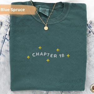 Peut inclure: Un sweat-shirt bleu épicéa avec le texte "CHAPTER 18" brodé en blanc, orné d'étoiles jaunes. Un collier doré avec un pendentif est posé sur le vêtement plié, sur un jean en denim.