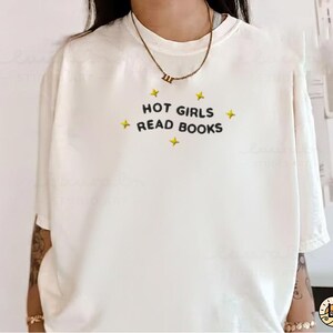 K&ouml;nnte beinhalten: Cremefarbenes T-Shirt mit dem schwarzen Schriftzug "HOT GIRLS READ BOOKS", umgeben von gelben Sternen. Das kurz&auml;rmlige Shirt hat eine lockere Passform und eignet sich f&uuml;r Freizeitkleidung.