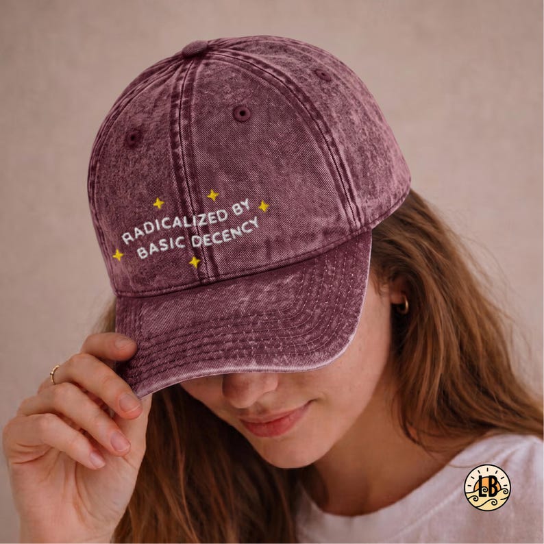 Op de afbeelding: Een bordeauxrode baseballpet met een acid-wash effect, voorzien van de geborduurde witte tekst "RADICALIZED BY BASIC DECENCY" in het midden, geaccentueerd door kleine gele ster-vormige iconen gestikt rondom de belettering op de stof.