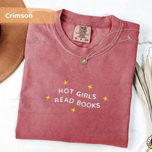 K&ouml;nnte beinhalten: Karmesinrotes T-Shirt mit der gestickten Aufschrift "HOT GIRLS READ BOOKS" und gelben Sternen. Das Shirt hat einen Rundhalsausschnitt und ein kleines Etikett am Kragen. Eine silberne Halskette mit Anh&auml;nger liegt auf dem Shirt.