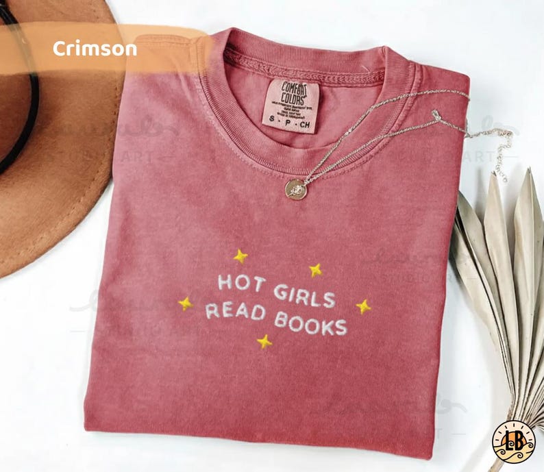 K&ouml;nnte beinhalten: Karmesinrotes T-Shirt mit der gestickten Aufschrift "HOT GIRLS READ BOOKS" und gelben Sternen. Das Shirt hat einen Rundhalsausschnitt und ein kleines Etikett am Kragen. Eine silberne Halskette mit Anh&auml;nger liegt auf dem Shirt.