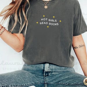 K&ouml;nnte beinhalten: Dunkelgraues T-Shirt mit dem wei&szlig;en Text "HOT GIRLS READ BOOKS", akzentuiert mit gelben Sternen. Das Shirt wird mit blauen Jeans getragen. Eine goldene Halskette und ein Armband sind ebenfalls sichtbar. L&auml;ssiger Stil.