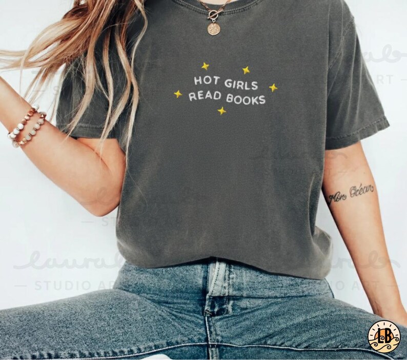 K&ouml;nnte beinhalten: Dunkelgraues T-Shirt mit dem wei&szlig;en Text "HOT GIRLS READ BOOKS", akzentuiert mit gelben Sternen. Das Shirt wird mit blauen Jeans getragen. Eine goldene Halskette und ein Armband sind ebenfalls sichtbar. L&auml;ssiger Stil.