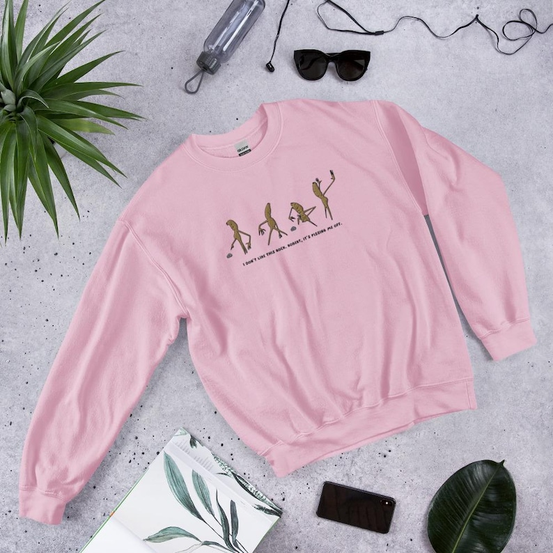 May include: A light pink sweatshirt with a graphic of stick figures in various poses, with the text "I DON'T LIKE THIS ROCK, MOMENT, IT'S PUSHING ME OFF." The sweatshirt is made of a soft material.
