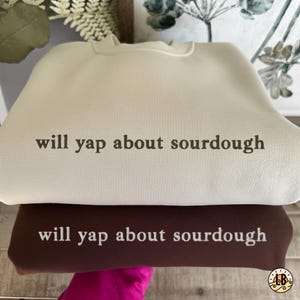 Könnte beinhalten: Zwei übereinander gestapelte Sweatshirts, eines cremefarben und eines braun. Beide haben den Aufdruck "will yap about sourdough" in einer kontrastierenden Farbe. Das cremefarbene Sweatshirt liegt oben.