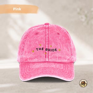 Op de afbeelding: Een felroze baseballpet met een acid-wash effect, voorzien van de geborduurde zwarte tekst THE BRIDE in het midden op het voorpaneel, omringd door kleine gele sterretjes. De pet heeft een gestructureerde kroon, een gebogen klep en een knoop.