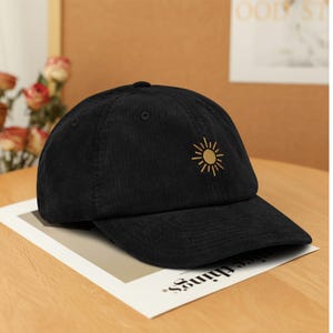 Könnte beinhalten: Schwarze Cord-Baseballkappe mit gebogenem Schirm. Auf der Vorderseite ist ein gelbes Sonnenmotiv aufgestickt. Die Kappe liegt auf einem Papier mit dem Aufdruck "nice things".
