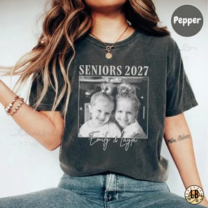 Könnte beinhalten: Ein anthrazitfarbenes Kurzarm-T-Shirt mit dem Text SENIORS 2027 über einem schwarz-weißen Foto von zwei lächelnden Kindern. Unter dem Foto sind die Namen Emily & Layla in einer weißen Schreibschrift auf der Vorderseite aufgedruckt.