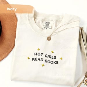 K&ouml;nnte beinhalten: Elfenbeinfarbenes T-Shirt mit dem Text "HOT GIRLS READ BOOKS" in Schwarz, umgeben von gelben Sternen. Eine goldene Halskette mit Anh&auml;nger liegt auf dem Shirt. Ein brauner Hut und eine getrocknete Pflanze sind im Hintergrund.