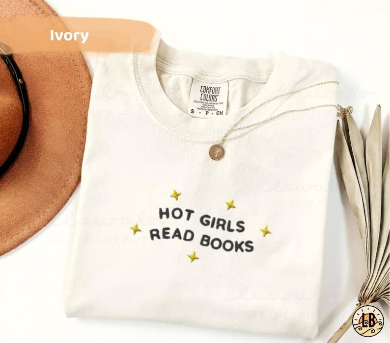 K&ouml;nnte beinhalten: Elfenbeinfarbenes T-Shirt mit dem Text "HOT GIRLS READ BOOKS" in Schwarz, umgeben von gelben Sternen. Eine goldene Halskette mit Anh&auml;nger liegt auf dem Shirt. Ein brauner Hut und eine getrocknete Pflanze sind im Hintergrund.