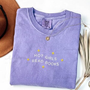K&ouml;nnte beinhalten: Ein lavendelfarbenes Comfort Colors T-Shirt mit dem gestickten Text "HOT GIRLS READ BOOKS" in Gelb. Eine goldene Halskette mit Anh&auml;nger liegt &uuml;ber dem Shirt. Das Shirt ist auf einer wei&szlig;en Oberfl&auml;che gefaltet.
