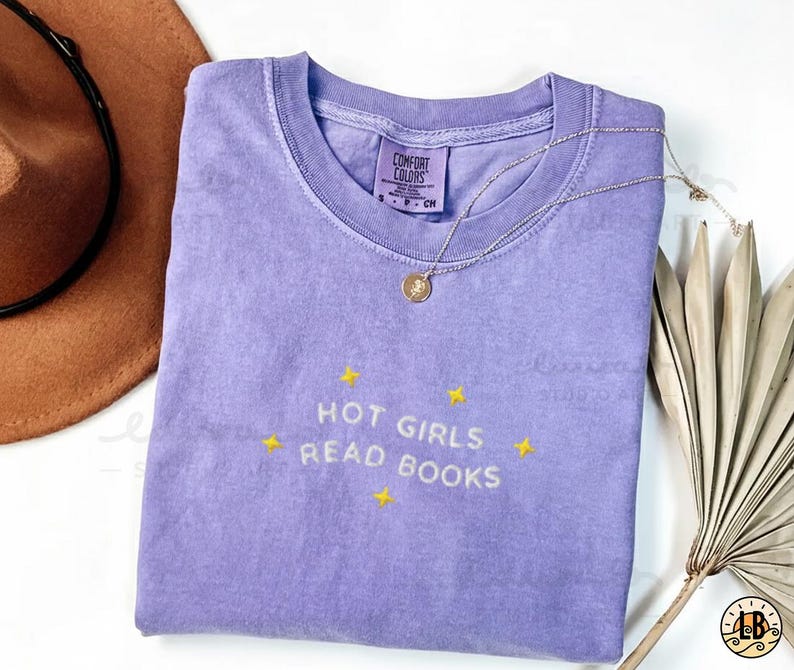 K&ouml;nnte beinhalten: Ein lavendelfarbenes Comfort Colors T-Shirt mit dem gestickten Text "HOT GIRLS READ BOOKS" in Gelb. Eine goldene Halskette mit Anh&auml;nger liegt &uuml;ber dem Shirt. Das Shirt ist auf einer wei&szlig;en Oberfl&auml;che gefaltet.