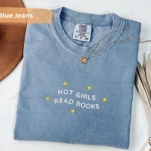 K&ouml;nnte beinhalten: Hellblaues T-Shirt mit dem gestickten Text "HOT GIRLS READ BOOKS" und gelben Sternen. Eine silberne Halskette mit Anh&auml;nger liegt auf dem Shirt. Das Shirt ist auf einer wei&szlig;en Oberfl&auml;che gefaltet.