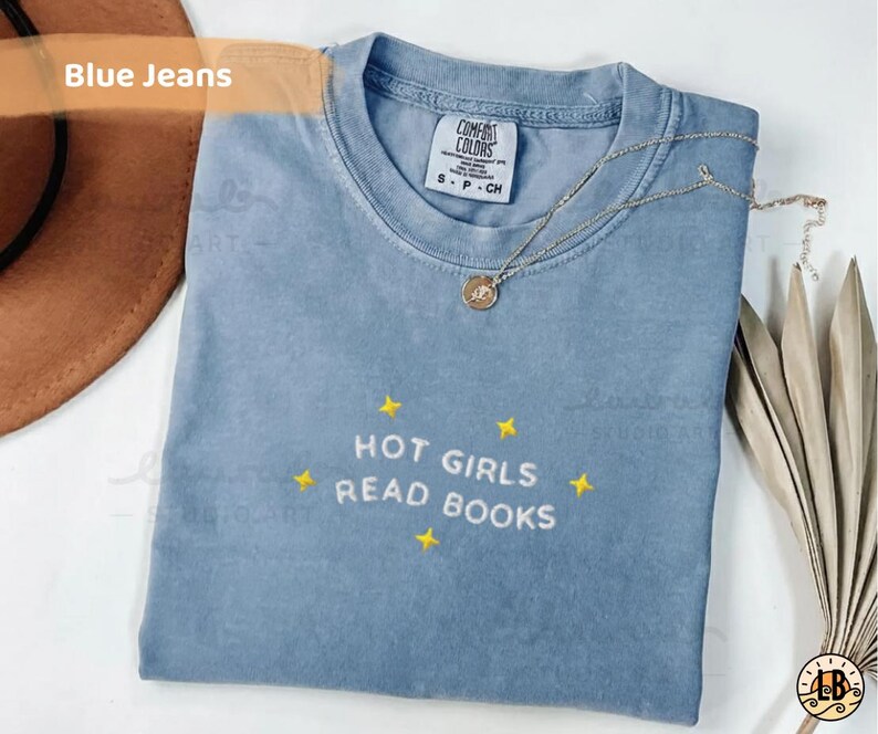 K&ouml;nnte beinhalten: Hellblaues T-Shirt mit dem gestickten Text "HOT GIRLS READ BOOKS" und gelben Sternen. Eine silberne Halskette mit Anh&auml;nger liegt auf dem Shirt. Das Shirt ist auf einer wei&szlig;en Oberfl&auml;che gefaltet.