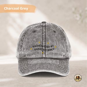 Op de afbeelding: Een antracietgrijze baseballpet met acid-wash effect en de geborduurde zwarte tekst "RADICALIZED BY BASIC DECENCY" in het midden van het voorpaneel, geaccentueerd door kleine gele stervormige iconen boven en onder de tekst.
