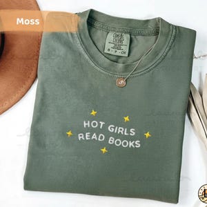 K&ouml;nnte beinhalten: Ein moosgr&uuml;nes T-Shirt mit dem gestickten Text "HOT GIRLS READ BOOKS" und gelben Sternen. Das Shirt ist gefaltet, mit einer goldenen Halskette und einem braunen Hut im Hintergrund.