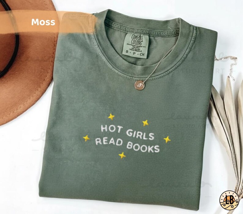 K&ouml;nnte beinhalten: Ein moosgr&uuml;nes T-Shirt mit dem gestickten Text "HOT GIRLS READ BOOKS" und gelben Sternen. Das Shirt ist gefaltet, mit einer goldenen Halskette und einem braunen Hut im Hintergrund.