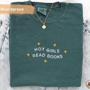 K&ouml;nnte beinhalten: Ein blau-fichtefarbenes T-Shirt mit dem gestickten Text "HOT GIRLS READ BOOKS" in Wei&szlig;, umgeben von gelben Sternen. Eine goldene Halskette mit Anh&auml;nger liegt &uuml;ber dem Shirt, das auf Jeans gefaltet ist.