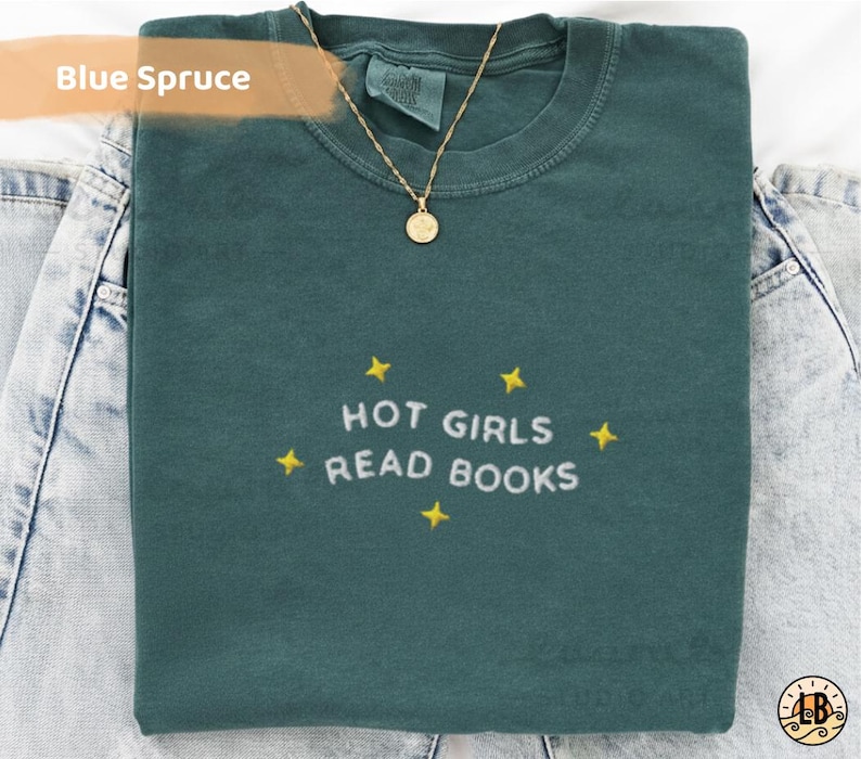 K&ouml;nnte beinhalten: Ein blau-fichtefarbenes T-Shirt mit dem gestickten Text "HOT GIRLS READ BOOKS" in Wei&szlig;, umgeben von gelben Sternen. Eine goldene Halskette mit Anh&auml;nger liegt &uuml;ber dem Shirt, das auf Jeans gefaltet ist.
