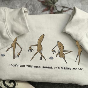 May include: Cream-colored sweatshirt with a humorous graphic of anthropomorphic root vegetables interacting with a small rock. The text "I DON'T LIKE THIS ROCK. ROBERT. IT'S PISSING ME OFF." is printed below the graphic. The sweatshirt has a crew neck and long sleeves.