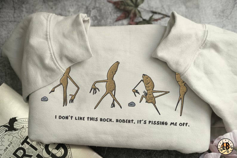 May include: Cream-colored sweatshirt with a humorous graphic of anthropomorphic root vegetables interacting with a small rock. The text "I DON'T LIKE THIS ROCK. ROBERT. IT'S PISSING ME OFF." is printed below the graphic. The sweatshirt has a crew neck and long sleeves.
