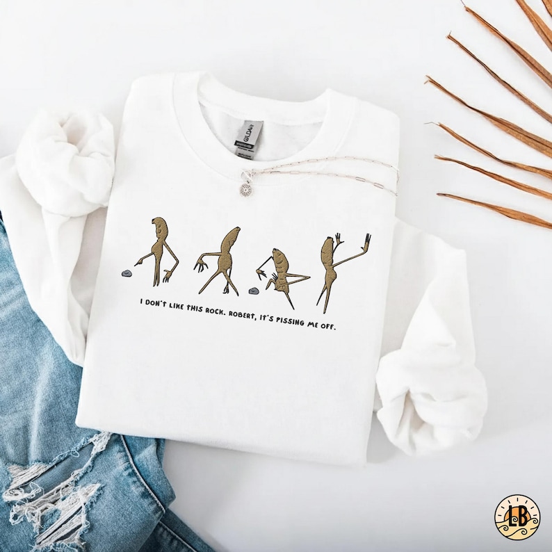 May include: White sweatshirt with a humorous graphic of four anthropomorphic figures with the text "I don't like this rock. Robert, it's pissing me off." A silver necklace with a pendant is draped on the sweatshirt.