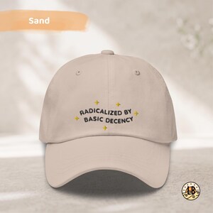 Op de afbeelding: Een zandkleurige baseballpet met de geborduurde zwarte tekst "RADICALIZED BY BASIC DECENCY" gecentreerd op het voorpaneel, geaccentueerd door vier kleine gele sterpictogrammen rondom de tekst, met een gebogen klep en ventilatiegaatjes.
