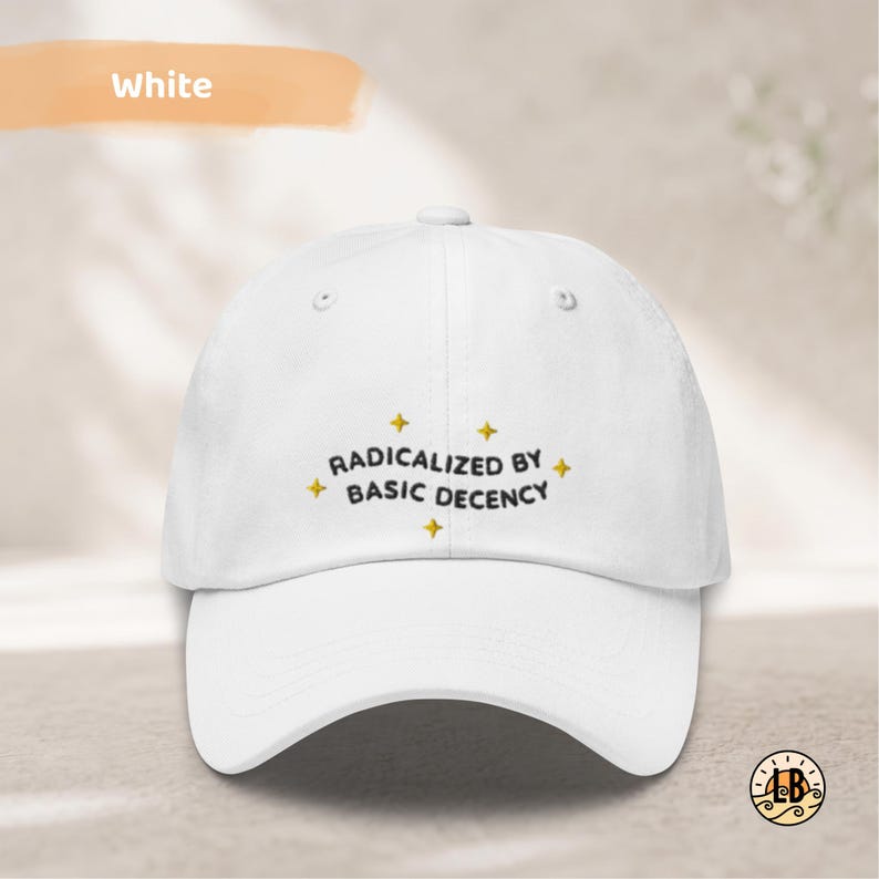 Op de afbeelding: Een witte baseballpet met zwarte geborduurde tekst die RADICALIZED BY BASIC DECENCY leest, geaccentueerd met kleine gele vierpuntige sterpictogrammen rondom de belettering. De pet heeft een gestructureerde kroon en een gebogen klep.