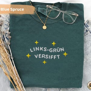 Könnte beinhalten: Ein gefaltetes, blau-fichtefarbenes T-Shirt mit dem Text "LINKS-GRÜN VERSIFFT" in Weiß mit gelben Sternen bestickt. Eine goldene Halskette mit Anhänger und eine Brille mit klarem Rahmen liegen obenauf. Das Shirt liegt auf einem Jeans-Hintergrund.