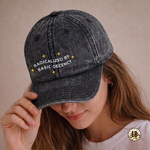 Op de afbeelding: Een antracietgrijze baseballpet met een acid-wash effect, voorzien van de geborduurde witte tekst "RADICALIZED BY BASIC DECENCY" op het voorpaneel, geaccentueerd door kleine gele sterretjes die boven en onder de tekst zijn gestikt.