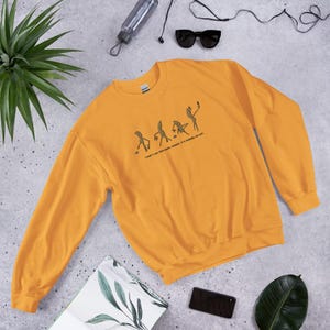 May include: Mustard yellow sweatshirt with a crew neck. The sweatshirt features a graphic of stick figures in various poses, with the text "I don't like this book, but it's pleasing me sex."