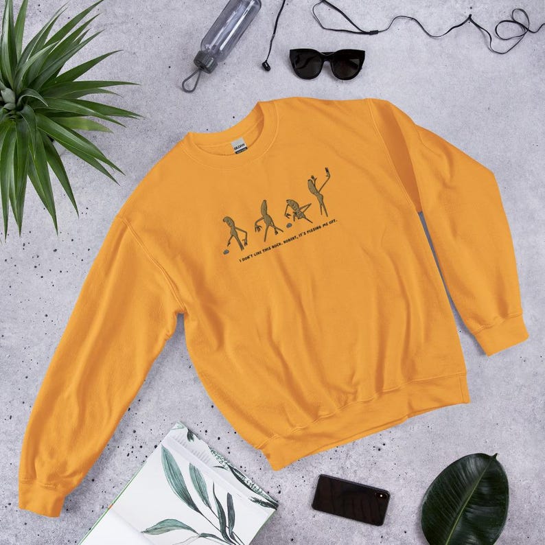 May include: Mustard yellow sweatshirt with a crew neck. The sweatshirt features a graphic of stick figures in various poses, with the text "I don't like this book, but it's pleasing me sex."