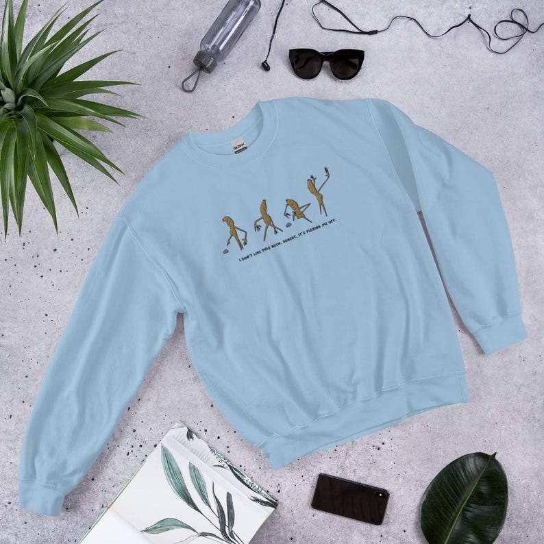 May include: A light blue sweatshirt with a humorous graphic of stick figures in various poses, with the text "I DON'T LIKE THIS ROCK, HONEST, IT'S POSING AS ART." The sweatshirt is made of a soft material.
