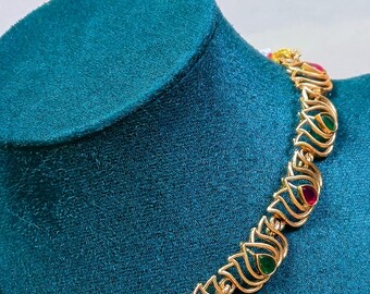 May include: A gold necklace with a lotus flower design and red and green gemstones. The necklace is on a blue velvet display stand. The earrings are also gold with a lotus flower design and red gemstones.