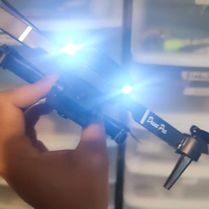 Puede incluir: Un dron negro con detalles blancos y el texto "Drone Pro" en el lateral. El dron tiene dos luces blancas brillantes en la parte delantera.