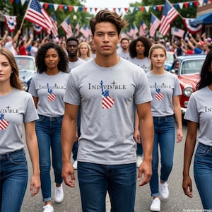 May include: A light grey short-sleeve t-shirt featuring the text INDIVISIBLE in black lettering, with the letter I replaced by a graphic of the Statue of Liberty wearing a crown and holding a torch, with a red, white, and blue flag pattern.