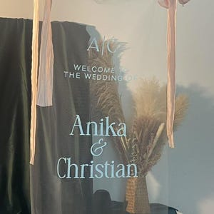 K&ouml;nnte beinhalten: Transparente Hochzeits-Willkommenstafel mit den Namen "Anika & Christian" und dem Datum "12.07.2025" in Wei&szlig;. Das Schild ist mit Trockenblumen verziert und mit rosa B&auml;ndern gebunden.