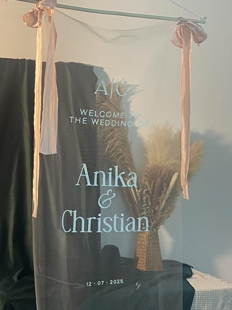 K&ouml;nnte beinhalten: Transparente Hochzeits-Willkommenstafel mit den Namen "Anika & Christian" und dem Datum "12.07.2025" in Wei&szlig;. Das Schild ist mit Trockenblumen verziert und mit rosa B&auml;ndern gebunden.
