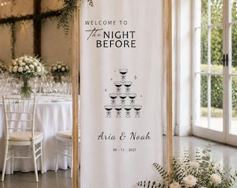 Może przedstawiać: Wysoki, pionowy baner z białej tkaniny zawieszony na jasnej drewnianej ramie. Baner zawiera czarny napis "WELCOME TO The NIGHT BEFORE", grafikę wieży z kieliszków do szampana, imiona "Aria & Noah" oraz datę "09 · 11 · 2027".