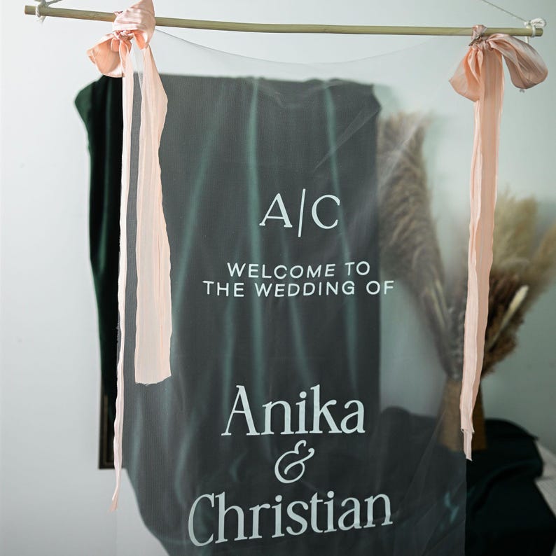 K&ouml;nnte beinhalten: Ein Hochzeits-Willkommensschild mit den Initialen "A/C" und dem Text "Welcome to the Wedding of Anika & Christian" in wei&szlig;er Schrift. Das Schild h&auml;ngt an einer Holzstange mit pfirsichfarbenen B&auml;ndern.