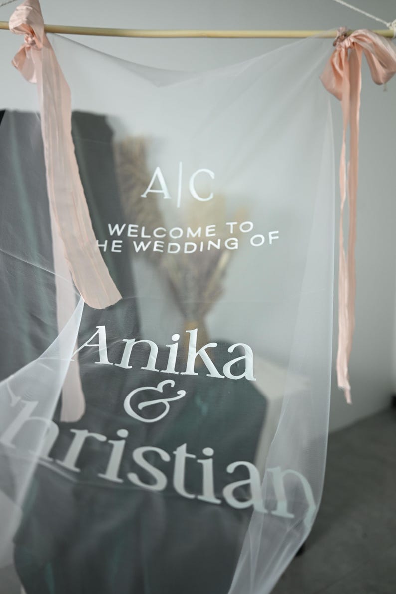 K&ouml;nnte beinhalten: Transparente Hochzeitsdekoration mit wei&szlig;em Text "Welcome to the Wedding of Anika & Christian". Das Schild ist mit rosa B&auml;ndern verziert und h&auml;ngt an einer Holzstange.