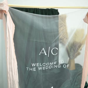 K&ouml;nnte beinhalten: Ein durchsichtiges, graues Hochzeitsbanner mit dem Text "A/C WELCOME TO THE WEDDING OF". Das Banner ist an einem Holzstab aufgeh&auml;ngt und mit pfirsichfarbenen B&auml;ndern und floralen Akzenten verziert.