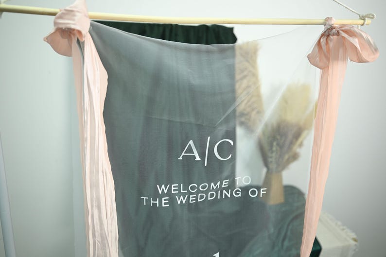 K&ouml;nnte beinhalten: Ein durchsichtiges, graues Hochzeitsbanner mit dem Text "A/C WELCOME TO THE WEDDING OF". Das Banner ist an einem Holzstab aufgeh&auml;ngt und mit pfirsichfarbenen B&auml;ndern und floralen Akzenten verziert.