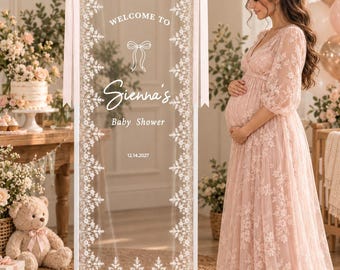 May include: A rectangular vertical sign with a white frame and a sheer center panel featuring white floral lace borders. The text reads Welcome to Sienna's Baby Shower 12.14.2027 with a small bow icon. Two pale pink ribbons are tied to the top corners.