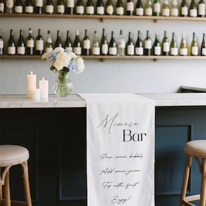 May include: A mimosa bar setup with a white runner displaying instructions. The bar features a dark blue base, marble countertop, and two stools. A vase of flowers and candles are on the counter. Shelves of wine bottles are in the background.