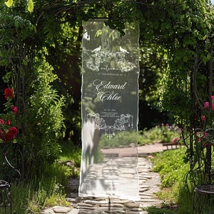 K&ouml;nnte beinhalten: Transparente Hochzeitsanzeige mit der Aufschrift "Welcome to the Wedding of Edward & Chloe" in eleganter Schrift. Das Schild ist mit dekorativen Verzierungen und Vogelsilhouetten versehen. Es befindet sich in einem Garten.