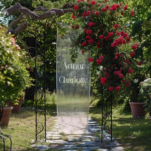 K&ouml;nnte beinhalten: Ein durchsichtiges Hochzeitszeichen aus Acryl mit den Namen "Arthur & Charlotte" in wei&szlig;er Schrift. Das Schild wird von einem schwarzen Schmiedeeisenbogen umrahmt, umgeben von Gr&uuml;n und roten Rosen. Das Hochzeitsdatum ist ebenfalls zu sehen.