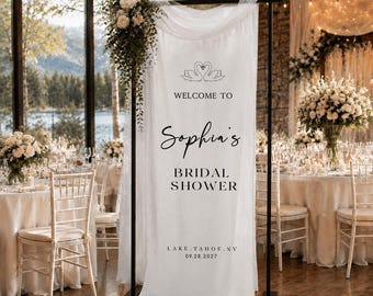 Może przedstawiać: Pionowy baner z białej tkaniny zawieszony na czarnym metalowym stojaku, zawierający napis "WELCOME TO Sophia's BRIDAL SHOWER LAKE TAHOE, NV 09.28.2027" oraz grafikę dwóch łabędzi tworzących kształt serca, ozdobiony białymi akcentami kwiatowymi.