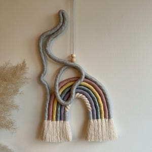 May include: A rainbow-colored macrame wall hanging with a gray rope design and wooden beads. The rainbow is made of cotton rope and has a fringe at the bottom.