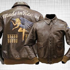 Op de afbeelding: Bruine leren bomberjack met een klassieke kraag en twee voorzakken. De achterkant van de jas heeft een geschilderd ontwerp met de woorden "Made in Heaven" en een gevleugelde pin-up girl. De jas heeft een geribbelde tailleband en manchetten.